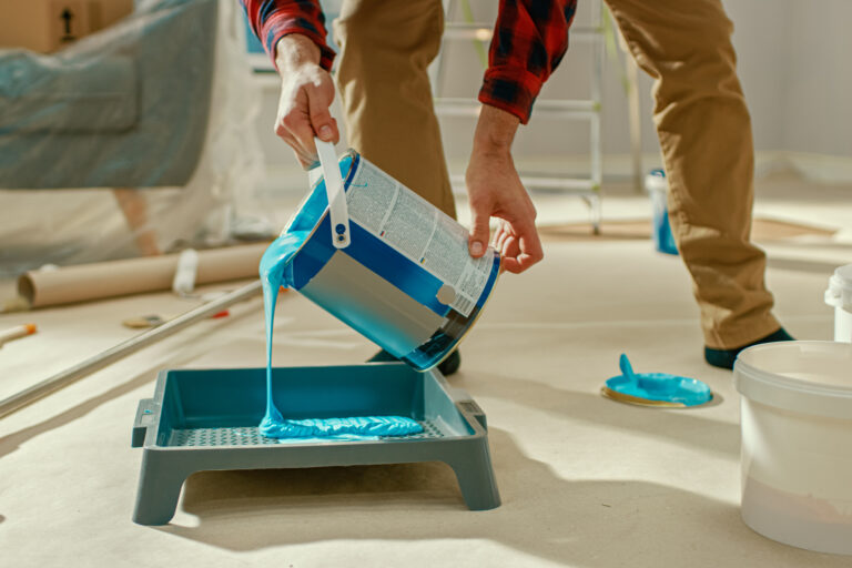 Young Man in Brown Jeans and Red Checked Shirt Picks Up a Can of Light Blue Paint and Pours it Into a Tray. Room Renovations at Home.